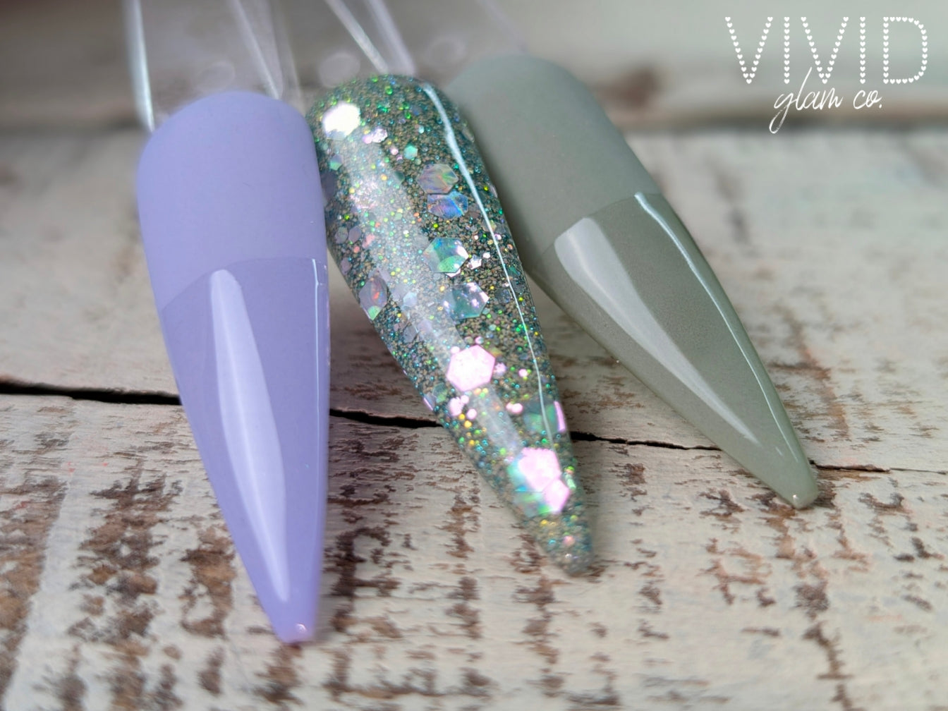Three nail dip powder samples in purple, hazel green,  glittery, and gray on a wooden surface with 'Vivid Glam Co' branding.