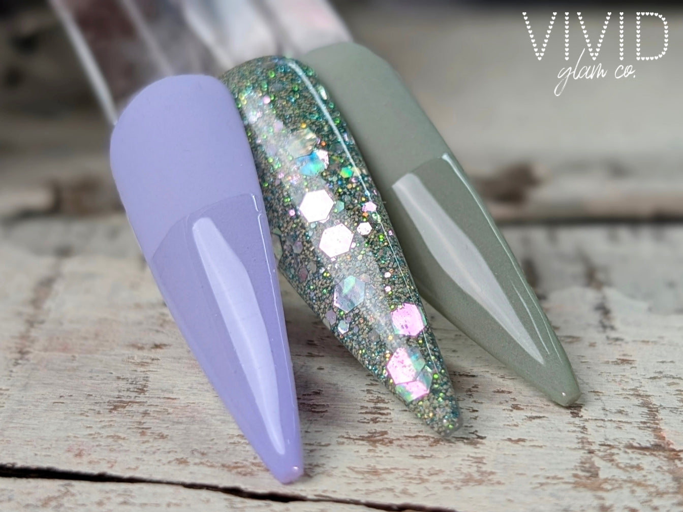 Nail art with glittery design on a wooden surface, featuring the brand 'Vivid glam co."