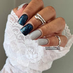 Vivid Glam Co manicure with a grey and navy blue glittery dip powder wearing a white lace sleeve.